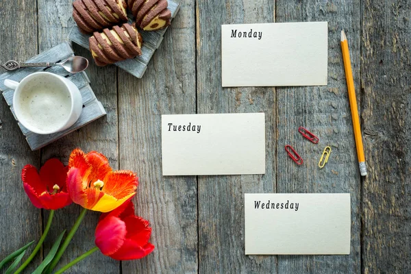 Tres tulipanes de colores sobre una mesa de madera. Una taza de café, galletas. Las hojas del diario Lunes Martes Miércoles — Foto de Stock