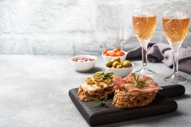 Tahta kesme tahtasında krem peynirli, pastırmalı ve zeytinli tam tahıllı sandviç. Bir bardak beyaz şarap. Boşluğu kopyalayan gri beton arkaplan.