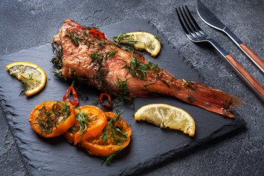 Fırınlanmış deniz levreği, domates biberli limon ve siyah tahta üzerinde siyah çimento arka planda duran otlar. Fırında pişirilmiş levrek balığı.