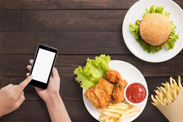 Usando smartphone com hambúrguer, batatas fritas e frango frito conjunto . — Fotografia de Stock