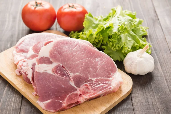 Raw pork on cutting board and vegetables on wooden background — Stock Photo, Image