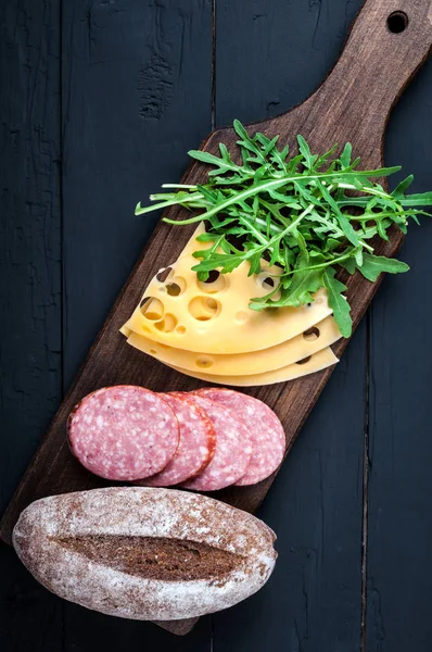 Madlavning sandwich. Ingredienser til sandwich pølse, maasdam ost, arugula salat og fuldkornsbrød på sort træ baggrund. Fødevarebaggrund - Stock-foto