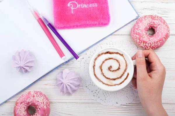 Espacio de trabajo femenino con taza de café, cuaderno de bocetos, bolígrafos, accesorios rosas y rosquillas. Mujer sosteniendo una taza de capuchino —  Fotos de Stock