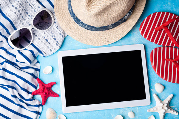 Summer women's accessories: sunglasses, hat, flip flop, shirt and tablet on blue background. Beach, vacations, travel and freelance