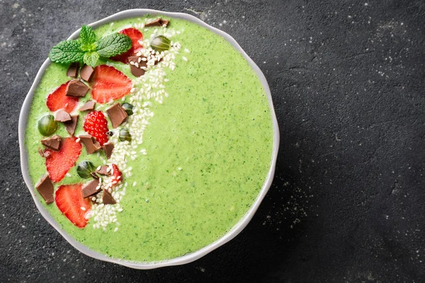 Ciotola di frullato verde con kiwi, spinaci, fragole, uva spina, cioccolato e semi di sesamo su sfondo di cemento scuro primo piano. Note di alimentazione, colazione e dieta. Copia spazio — Foto Stock