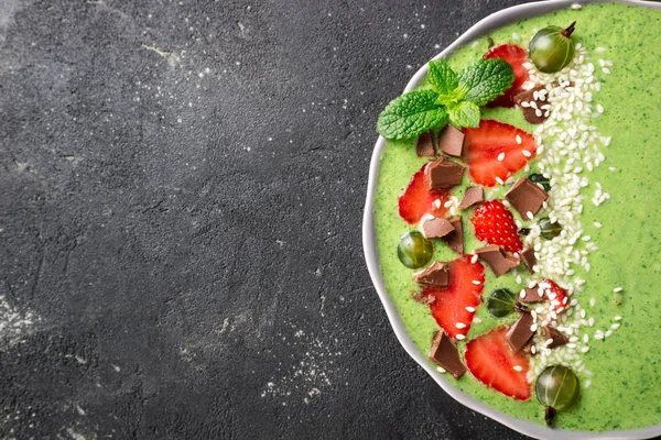 Ciotola di frullato verde con kiwi, spinaci, fragole, uva spina, cioccolato e semi di sesamo su sfondo di cemento scuro primo piano. Vista dall'alto — Foto Stock