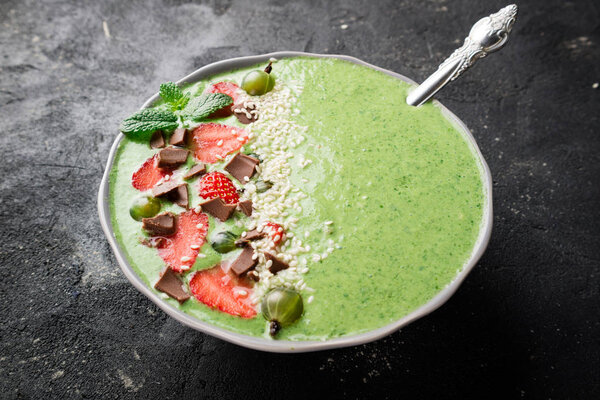 Breakfast smoothie bowl with kiwi, spinach, strawberry, gooseberry, chocolate and sesame seeds on dark concrete background. Green smoothie. Healthy eating diet