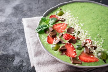 Kivi, ıspanak, çilek, bektaşi üzümü, çikolata ve susam tohumu karanlık beton zemin üzerine yeşil smoothie kase