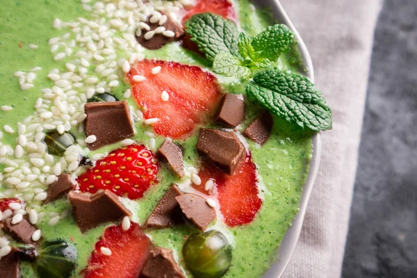 Smoothie bowl with kiwi, spinach, strawberry, gooseberry, chocolate and sesame seeds on dark concrete background closeup. Healthy eating, breakfast and diet concept. Top view — Stock Photo, Image