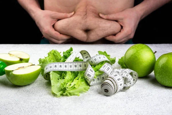 Man with fat belly and table with healthy eating. Dieting, slimming, weight loss and meal planning. Green apple, lettuce salad, glass of water, measuring tape