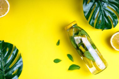 Monstera yaprakları ve Sarı zemin üzerine tropikal su şişesi. Detoks meyve suyu, narenciye ve nane yaprakları infüzyon. Üstten Görünüm, düz yatıyordu