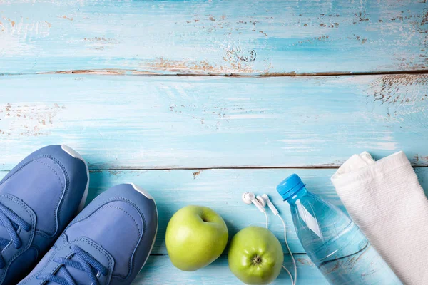 Plochá laických sportovní boty, láhev vody, jablka, ručník a sluchátka na modré dřevěné pozadí. Zdravý životní styl, sport a dietní koncepce — Stock fotografie