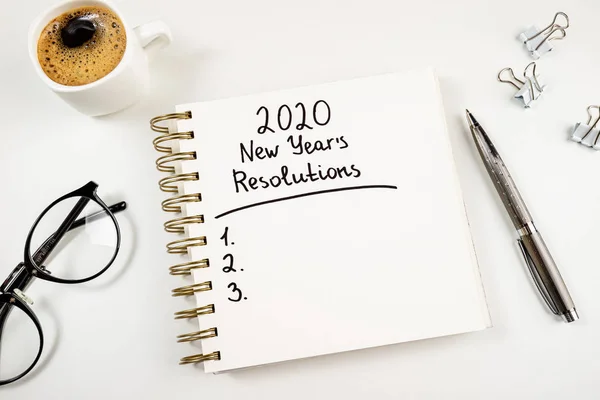 New year resolutions 2020 on desk. 2020 goals list with notebook, coffee cup and glasses on white background. Goal, plan, strategy, change, idea concept