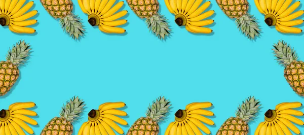 Colorful fruit pattern of fresh yellow bananas and pineapples on blue background. Frame of tropics fruit. Minimal summer food concept. Long format, top view, copy space