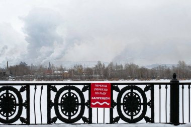 Nehir kenarındaki çitte yapılan duyuru. Mesaj çite sabitlendi. Kelimeler, geçiş, buzda hareket etmek yasaktır. Nehir manzarası, fabrika, baca ve duman..