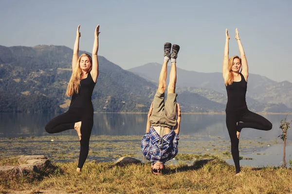 Young yogis doing yoga headstand and standing asana. Beautiful mountain lake landscape background. Complex amazing and funny yoga picture