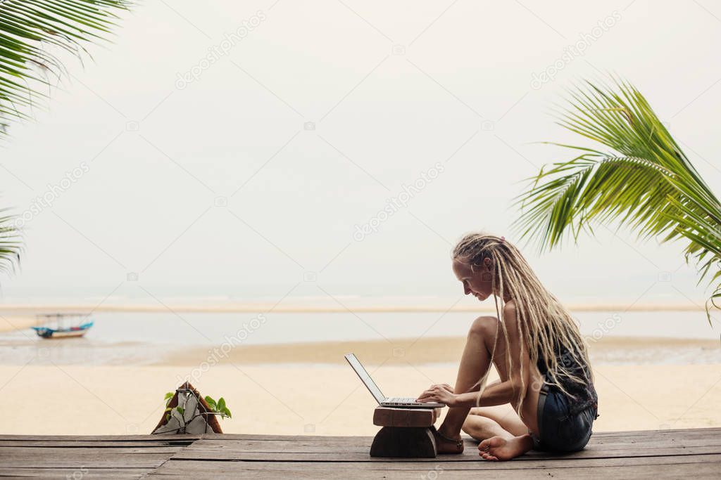 Young female wearing dreadlocks using laptop on sea shore. Shopping online, freelance, working outdoor