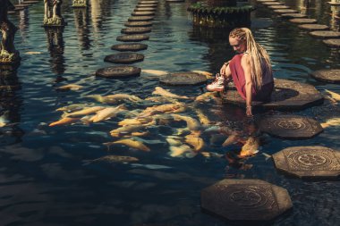 Woman feeding beautiful coloured fish in pond. Taman Tirtagangga temple on Bali clipart