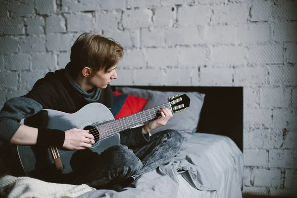 Genç Erkek Gitarist Yatakta Oturup Gitar Çalıyor Arkaplanda Beyaz Tuğla — Stok fotoğraf