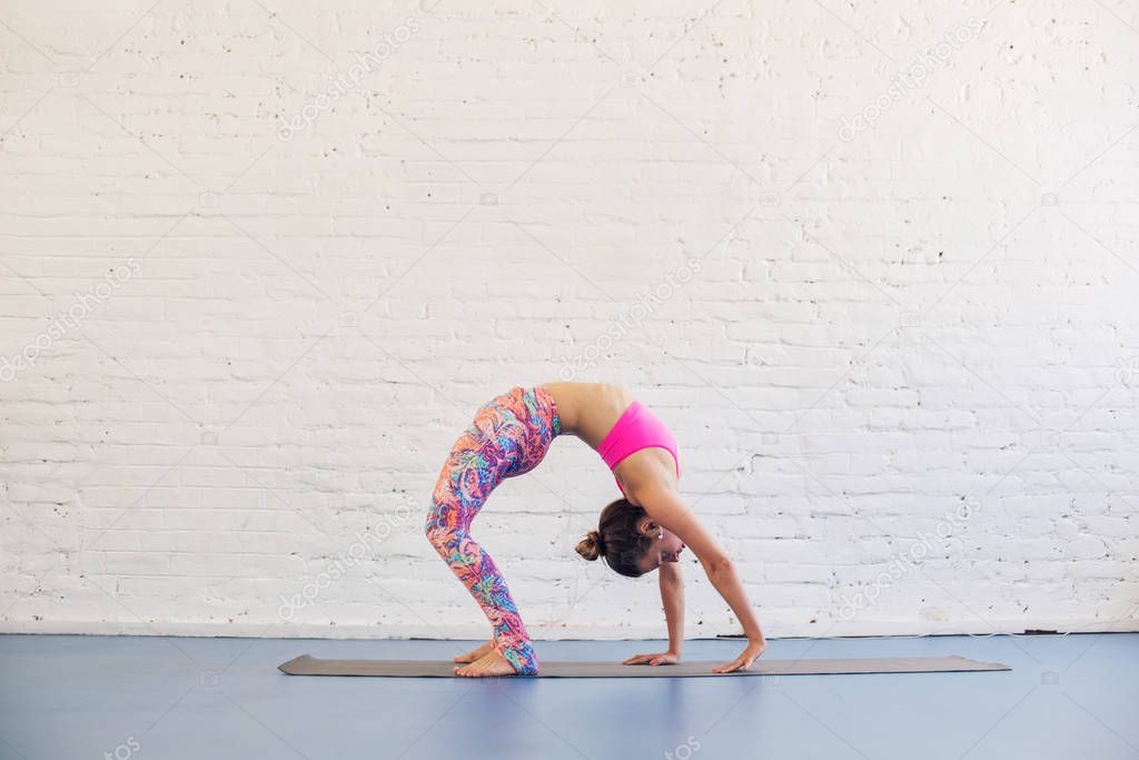 Flexible woman doing yoga exercise in studio. White brick wall on background 