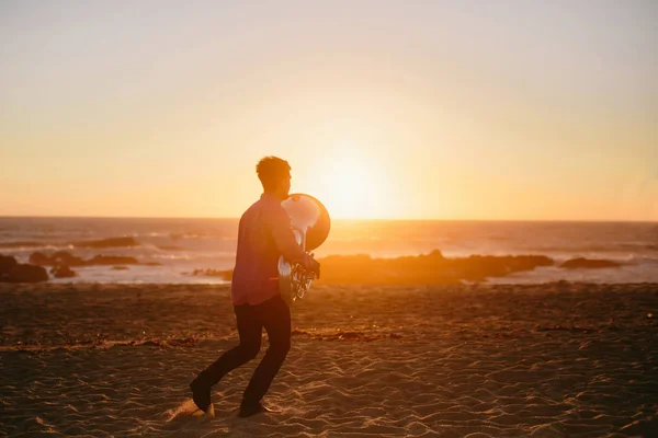 Silhouette of young man running with Tuba musical instrument  on rocky ocean coast during sunset. Music outdoor
