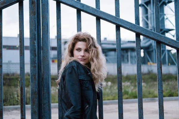 漂亮的女性穿着皮夹克站在室外 铁栅栏和背景工业景观 — 图库照片