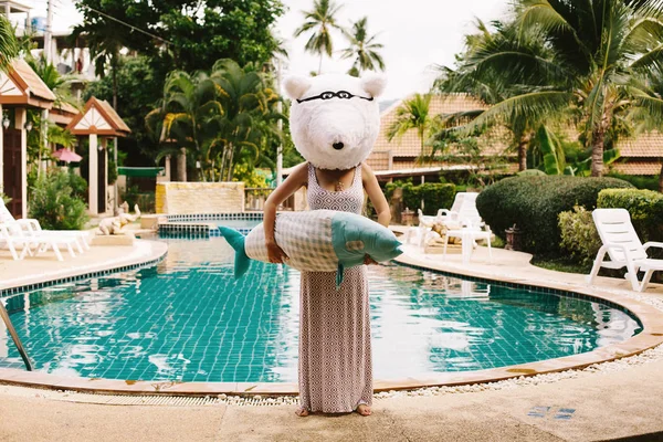 Funny woman dressed as a polar bear holding a big fish staying near swimming pool and palm trees. Christmas in asian tropics, hot summer day