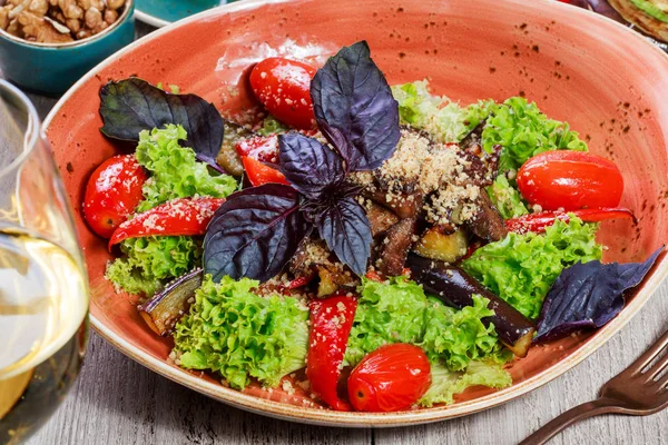 Ensalada caliente con tomates a la parrilla, berenjena, calabacín, pimiento rojo, hojas de ensalada, adornada con nueces ralladas y albahaca y copa de vino sobre fondo de madera. Comida italiana. Vista superior — Foto de Stock