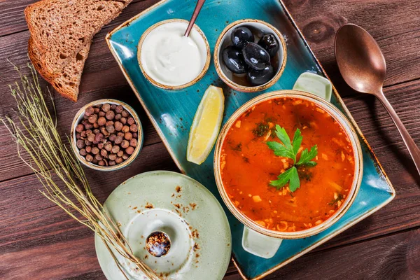 Zuppa con carne, olive, erbe aromatiche, limone, panna acida in ciotola, pane nero e spezie su fondo di legno scuro, cibo fatto in casa. Minestra russa tradizionale - solyanka. Vista dall'alto — Foto Stock