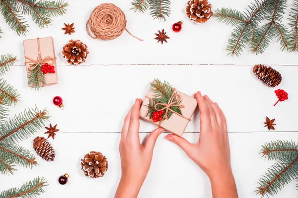 Fondo natalizio con rami di abete, pigne, matassa di iuta, decorazioni rosse. Mani femminili che tengono il regalo di Natale. Natale e Felice Anno Nuovo composizione. Posa piatta, vista dall'alto — Foto Stock