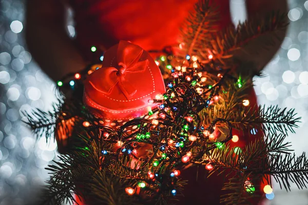 Mãos femininas segurando presente de Natal, ramo de abeto e decorações de luz multicoloridas, no fundo escuro do feriado . — Fotografia de Stock