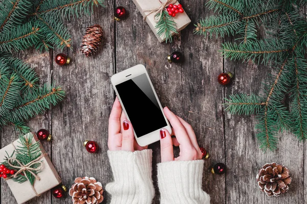 Mãos femininas segurando telefone celular no fundo de madeira com presentes de Natal, ramos de abeto. Composição de Natal e Feliz Ano Novo. Deitado plano, vista superior — Fotografia de Stock