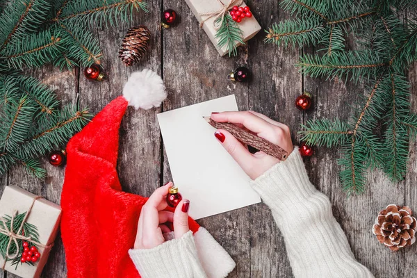 Mano femenina escribiendo una carta a Santa sobre fondo de madera con regalos de Navidad, ramas de abeto, conos de pino. Navidad y Feliz Año Nuevo composición. Piso tendido, vista superior — Foto de Stock