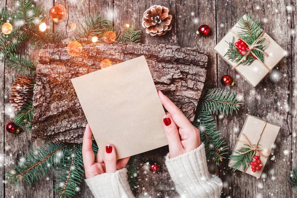 Mano femenina escribiendo una carta a Santa sobre fondo de madera con regalos de Navidad, textura de corteza, ramas de abeto, conos de pino, decoraciones rojas. Navidad y Feliz Año Nuevo composición. Piso tendido, vista superior — Foto de Stock