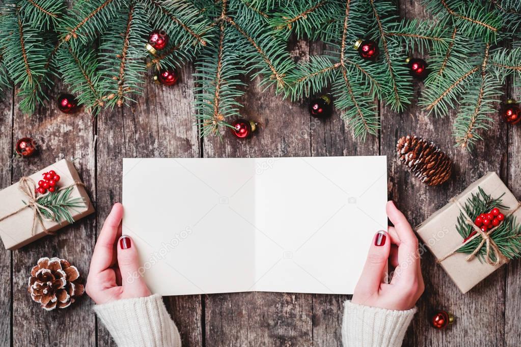 Female hand writing a letter to Santa on wooden background with Christmas gifts, bark texture, Fir branches, pine cones, red decorations. Xmas and Happy New Year card. Flat lay, top view
