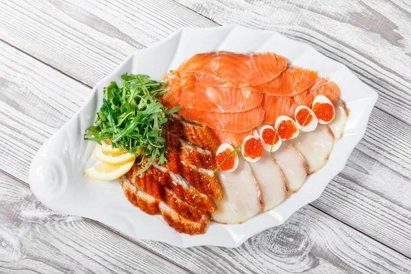 Seafood platter with salmon slice, smoke sturgeon, quail eggs with red caviar, slices fish fillet, decorated with arugula and lemon on wooden background close up. Mediterranean appetizers. Top view