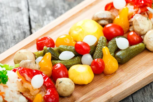 Assorted pickled vegetables - Sauerkraut cabbage, peppers, cucumbers, tomatoes, onions, mushrooms and herbs on cutting board on wooden background close up. Snacks and Wine appetizers set. Top view — Stock Photo, Image