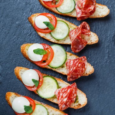 Caprese sandviç domates, mozzarella peyniri, fesleğen, salam taş arduvaz arka plan üzerinde ciabatta ekmeği üzerine kapatın. Lezzetli meze aperatifler, bruschetta, crostini, antipasti. Üstten Görünüm