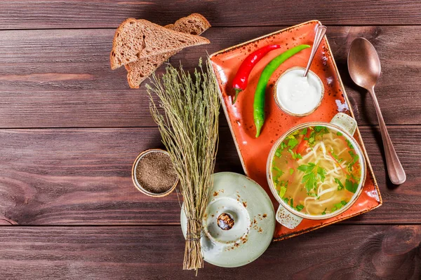 Vegetable soup, broth with noodles, herbs, parsley and vegetables in bowl with sour cream, spice, pepper, dried thyme and bread on dark wooden background. Ingredients on table. Top view — Stock Photo, Image