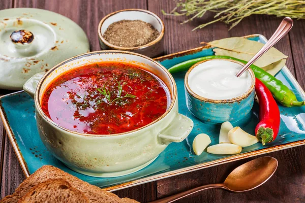 Sopa de remolacha tradicional ucraniana y rusa: borscht en maceta de barro con crema agria, especias, ajo, pimienta, hierbas secas y pan sobre fondo de madera oscura, comida saludable. Ingredientes en la mesa . — Foto de Stock