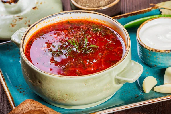 Sopa de remolacha tradicional ucraniana y rusa: borscht en maceta de barro con crema agria, especias, ajo, pimienta, hierbas secas y pan sobre fondo de madera oscura, comida saludable. Ingredientes en la mesa . — Foto de Stock