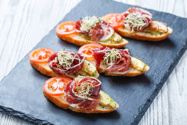 Appetizer bruschetta with prosciutto, tomato, zucchini on ciabatta bread on stone slate background close up. Delicious snacks, sandwiches, crostini, antipasti on party or picnic time. Top view