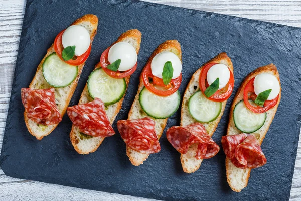 Sanduíches Caprese com tomate, queijo mussarela, manjericão, salame no pão ciabatta no fundo de ardósia de pedra perto. Deliciosos petiscos aperitivos, bruschetta, crostini, antipasti. Vista superior — Fotografia de Stock