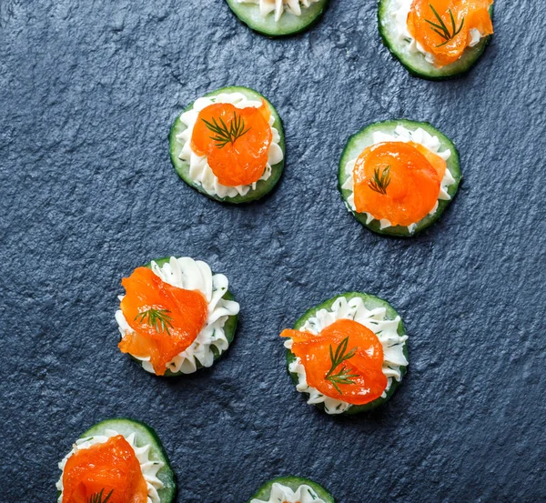 Appetizer canape with salmon, cucumber and cream cheese on stone slate background close up. Delicious snacks, sandwiches, crostini, bruschetta, antipasti on party or picnic time. Top view