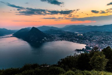 Dağlar ve akşam şehir Lugano dramatik günbatımı sırasında İsviçre Alpleri üzerinde çevrili lake Lugano hava görünümünü. Seyahat