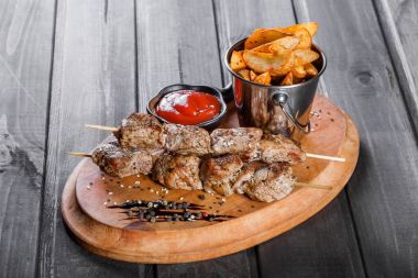 Skewers of Meat with sauce and potatoes fries in a bucket on wooden cutting board clipart