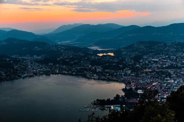 Vista aérea do lago Lugano cercado por montanhas e cidade noturna Lugano em durante o pôr do sol dramático, Suíça, Alpes. Viagens — Fotografia de Stock