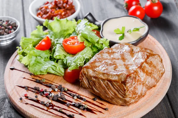 Vepřový grilovaný steak s zeleninový salát, rajčaty a omáčkou na dřevěném prkénku — Stock fotografie