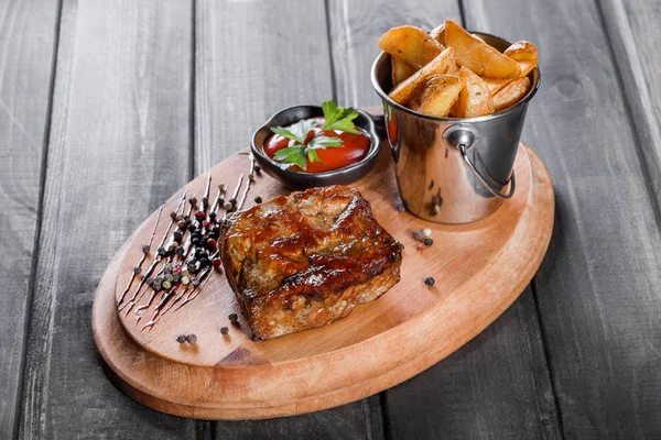 Bife grelhado com molho e batatas fritas em um balde na tábua de corte de madeira — Fotografia de Stock