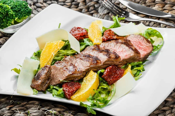 Grilled beef with orange fruit, arugula, parmesan cheese and sun-dried tomatoes on bamboo background. Traditional meat for Holidays. Top view